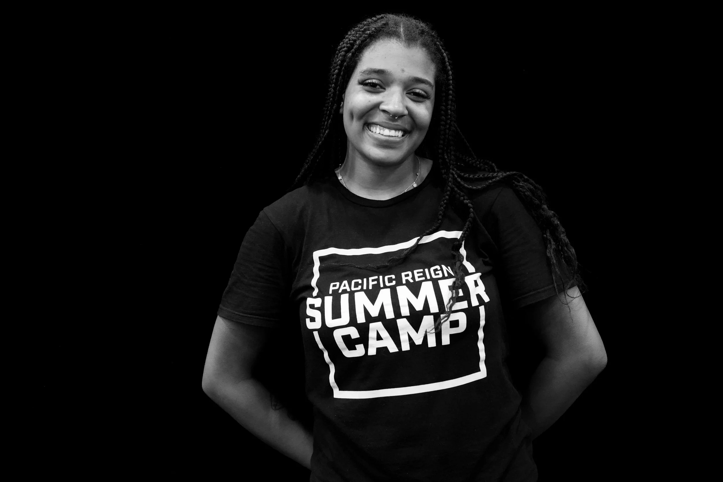 Black and white photo of a smiling woman with curly hair, wearing a t-shirt that reads 'Pacific Reign Gyms' against a dark background.