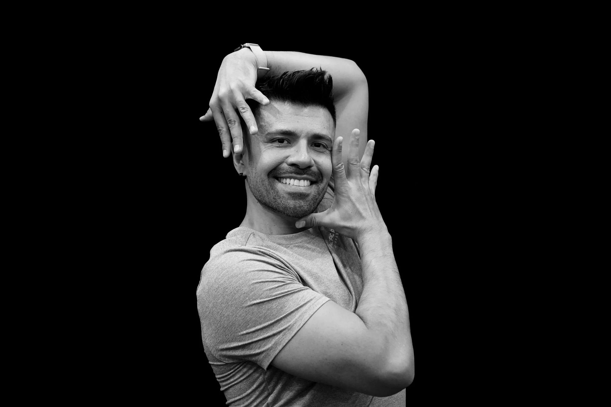 A young man with short hair and a beard poses with his hands framing his face and head, against a black background.