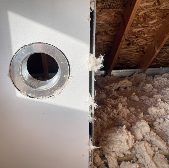 Metal pipe fitting in white drywall with wooden beams and insulation visible through opening.