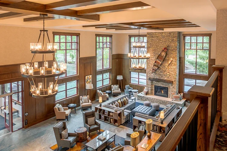 A spacious living room with wooden wall panels, large windows, a stone fireplace with a canoe and paddle decor, multiple chandeliers, and comfortable seating such as armchairs and sofas.