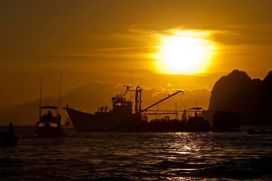 Silhouettes of boats on water during sunset with a large illuminated sun and rocky cliffs in the background.