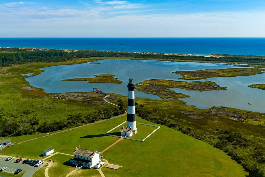 A lighthouse surrounded by grassy land, with water bodies and marshlands in the background, and the ocean beyond.