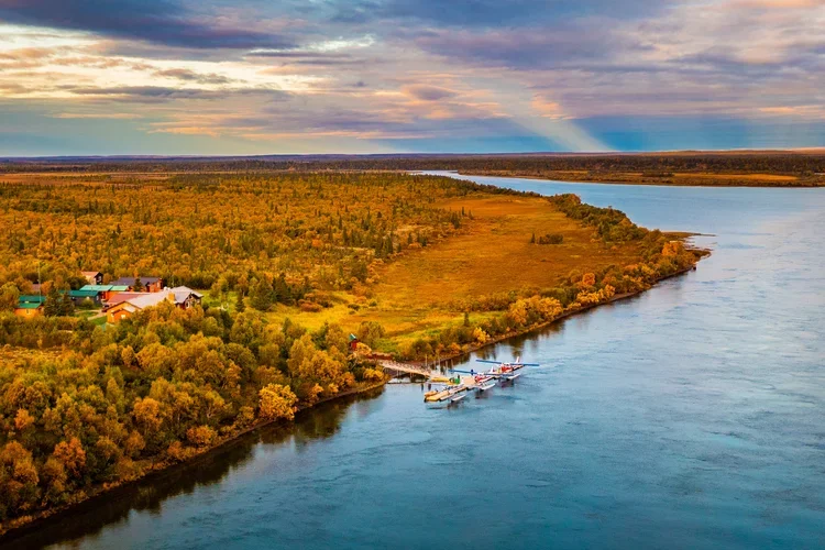 An aerial view of a river with a small airplane parked at a dock, surrounded by autumn-colored trees and a few houses under a partly cloudy sky.