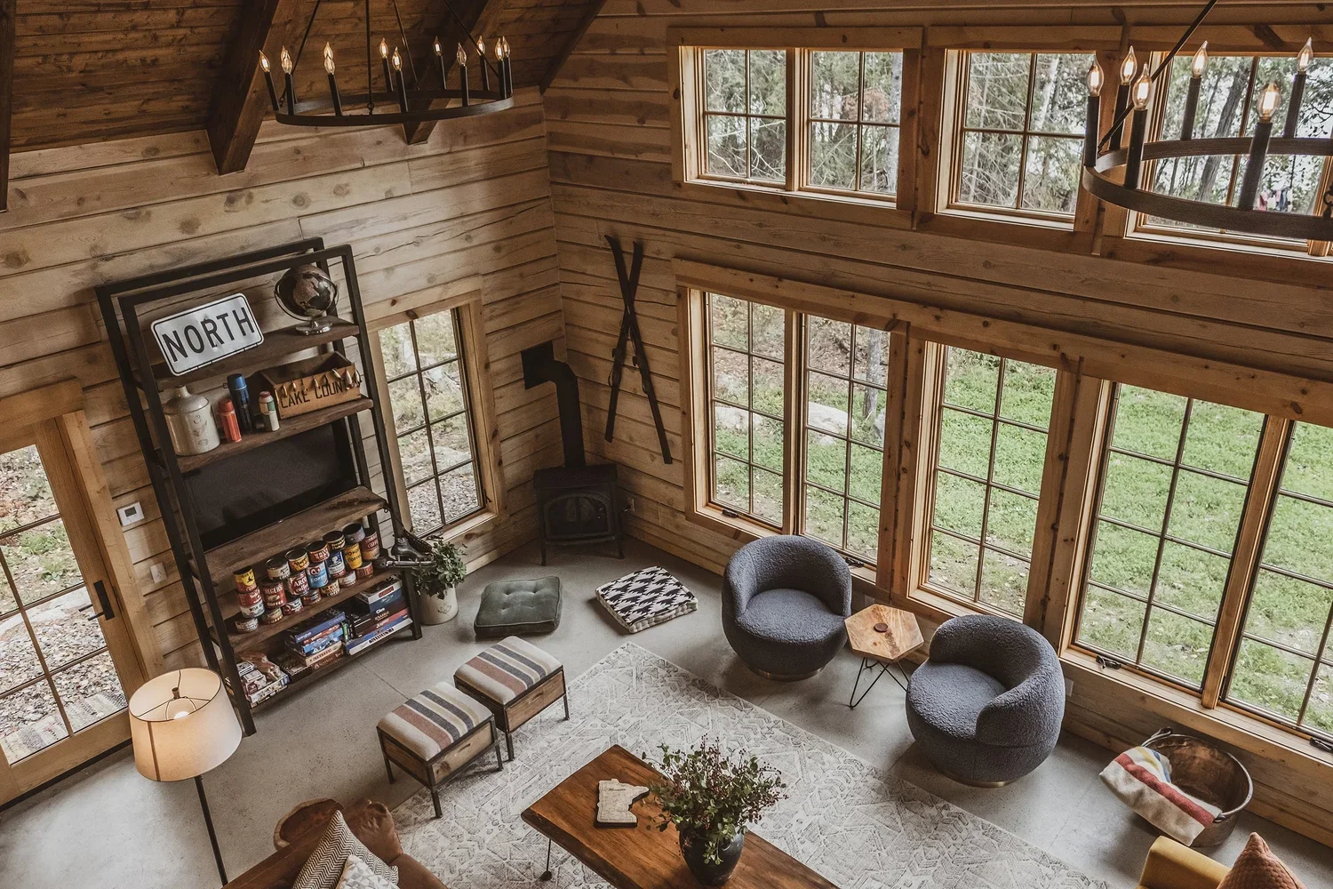 Living room with wood-paneled walls and large windows overlooking a green outdoor area, featuring two grey modern armchairs, a small wooden side table, a wooden coffee table with a vase and books, a patterned cushion, a wood stove with a black pipe, a black wall-mounted shelf with various books and decorative items, two small striped stools, a floor lamp, and a patterned rug.