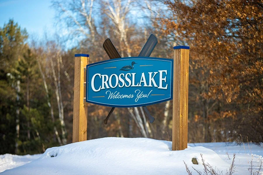 A welcome sign for Crosslake with cross-country skis behind it, set against a winter landscape with snow and trees with autumn foliage.