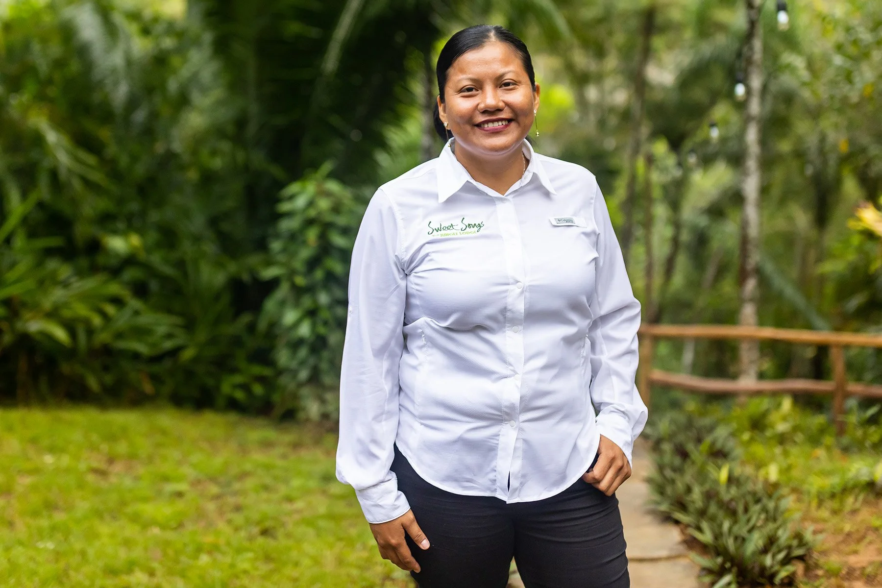 A woman smiling outdoors in a lush green park, wearing a white shirt with green embroidery and black pants.