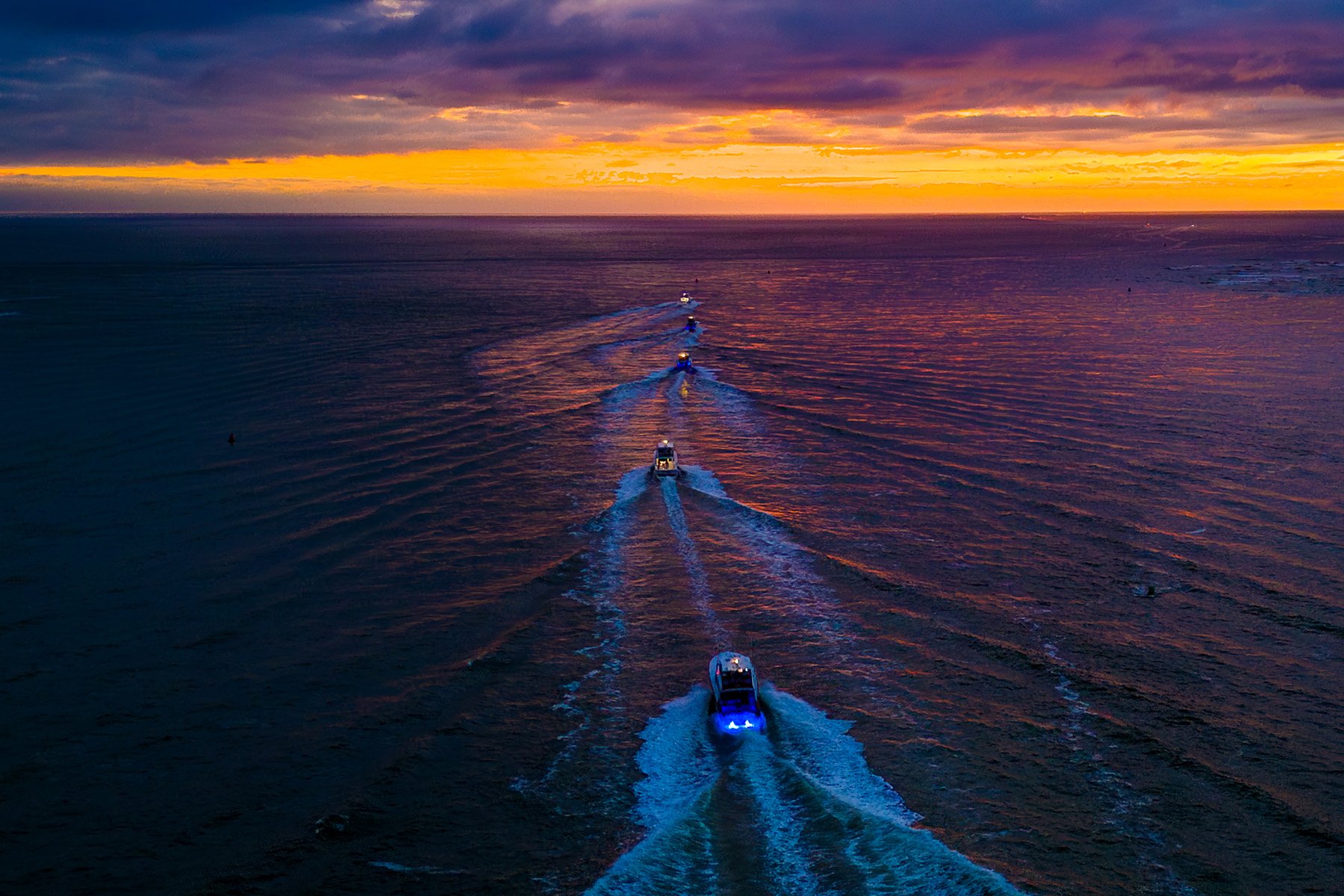 Multiple boats traveling in a line on the ocean during sunset, with vibrant orange, yellow, and purple clouds reflected on the water.