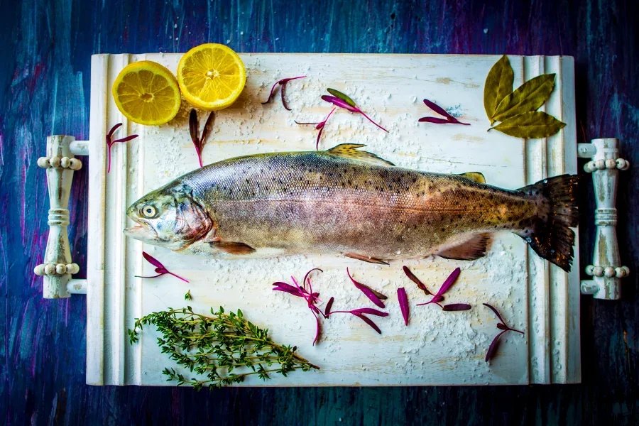 Fresh fish lying on a bed of flour or salt on a white cutting board, garnished with lemon slices, purple flower petals, bay leaves, and thyme, with a rustic blue wooden background.