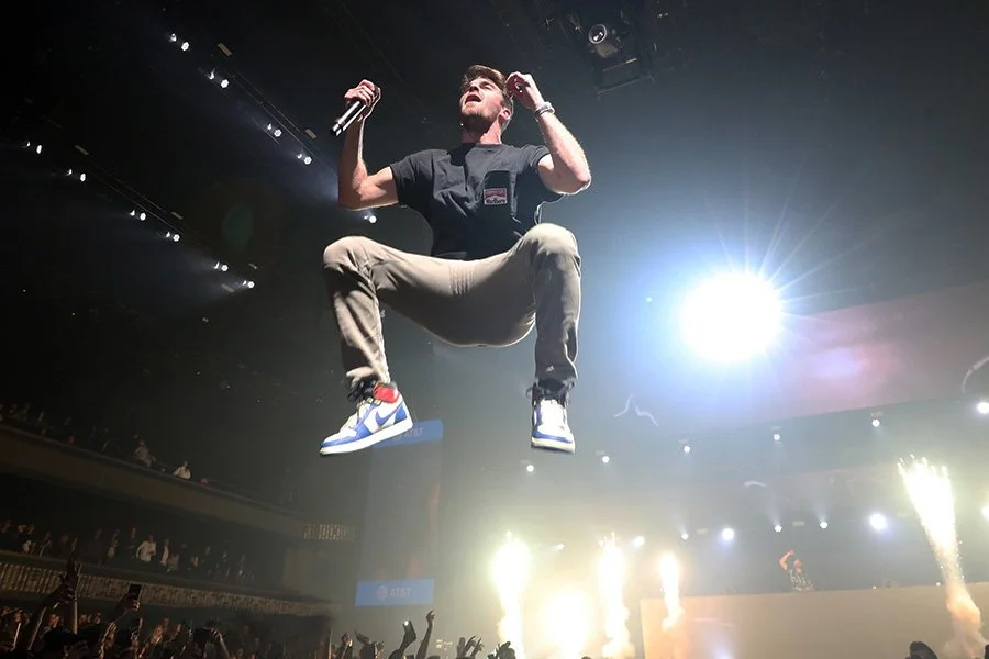 A man performing a leap on stage during a concert, holding a microphone, with bright stage lights and pyrotechnics in the background.