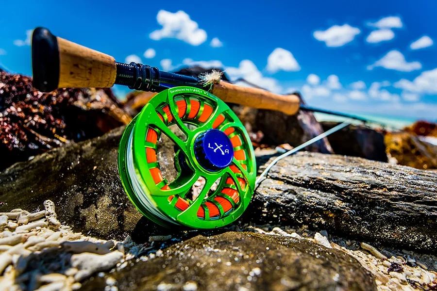 A green and orange fishing reel resting on rocks at a beach, with a fishing rod extended behind it and a partly cloudy blue sky in the background.