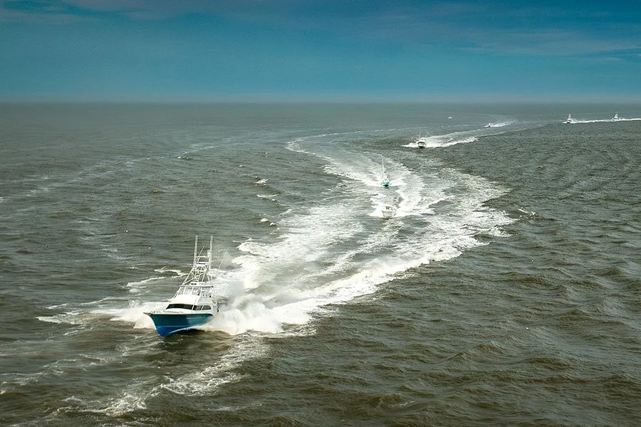 Multiple boats speed across open water, creating wakes and spray, with a clear sky overhead.
