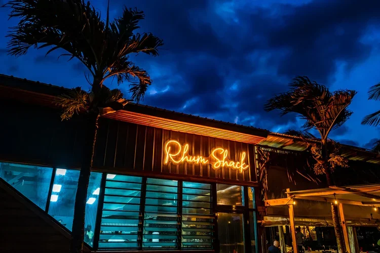 Nighttime view of the facade of Rhum Shack, a restaurant with large glass windows showing interior lighting, two palm trees in front, and a neon sign with the restaurant's name, under a dark blue sky with clouds.