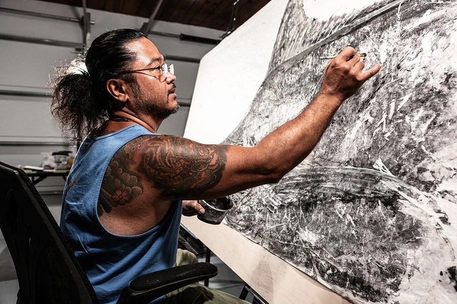 A man with long dark hair tied back, glasses, and tattoos on his arm, is painting a large, monochromatic abstract artwork on a canvas in a garage or studio.