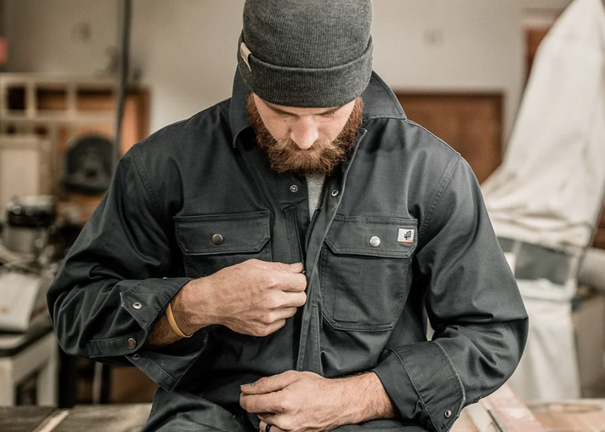 A man with a beard wearing a black jacket and a gray beanie, adjusting his jacket in a workshop.