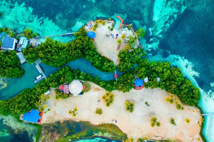 Aerial view of a small tropical island with sandy beaches, lush green trees, colorful buildings, and clear blue water surrounding it.