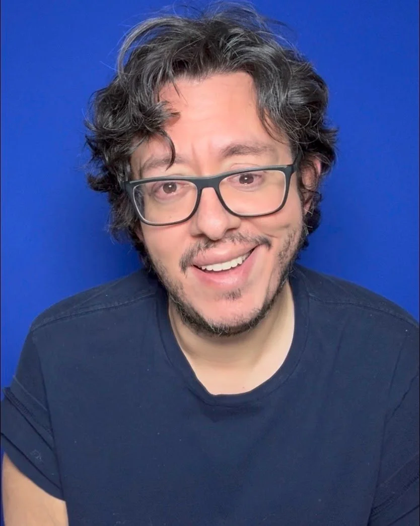 A man with glasses and curly hair smiling against a blue background.
