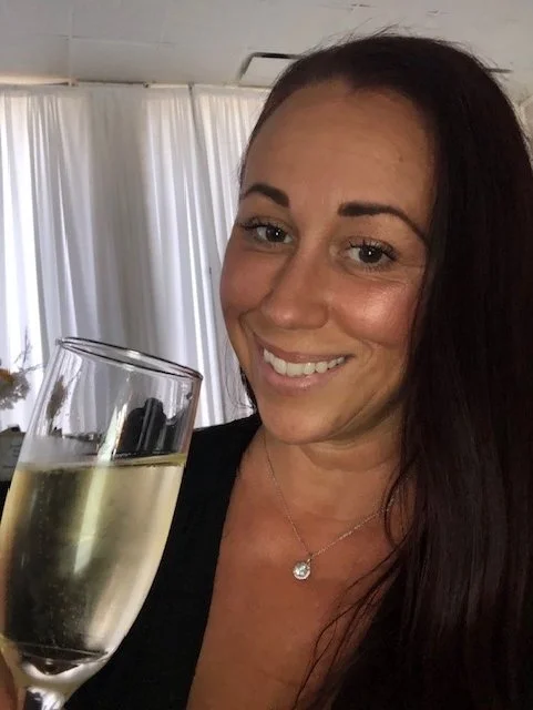 A woman smiling and holding a glass of champagne in a well-lit room with white curtains.