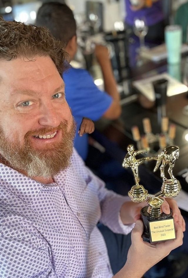 A man with curly hair and a beard smiling and holding a small golden trophy with a horse figure on top, which has a plaque reading "Best Blind Taster, The Unusual Suspects, 2023."