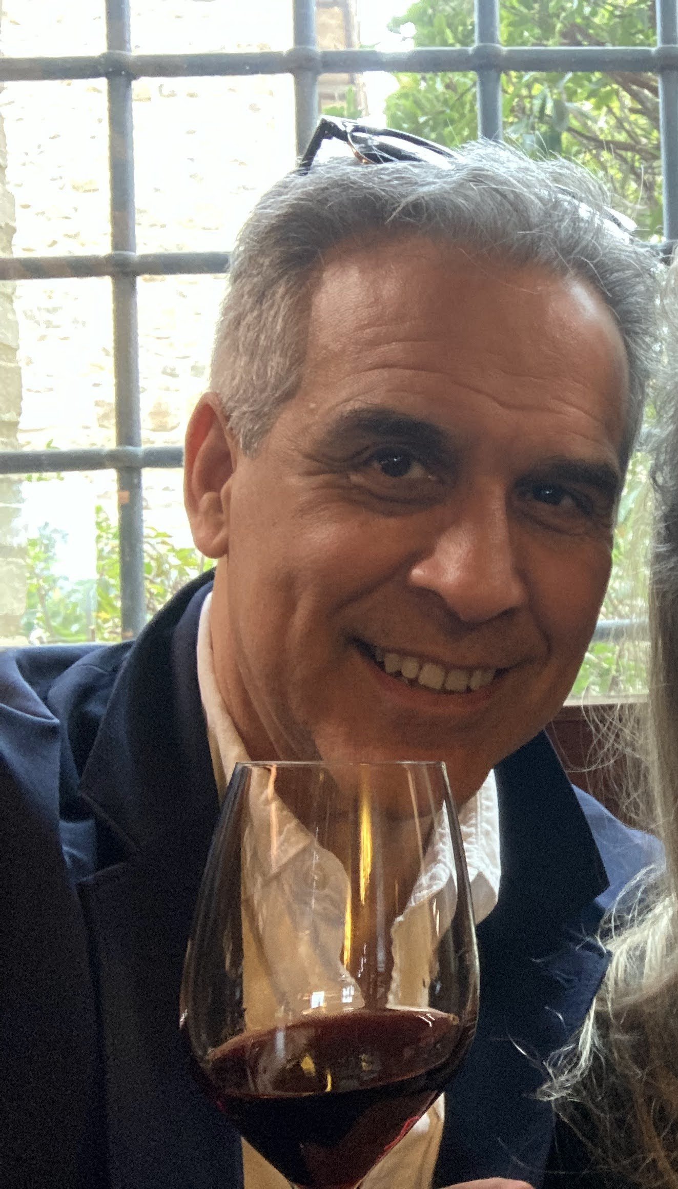 A smiling man holding a glass of red wine at a social gathering, with greenery visible through a window in the background.