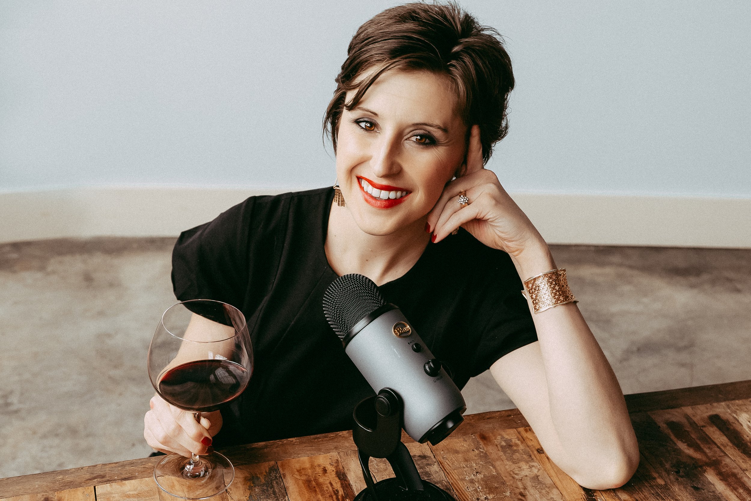 A woman with short brown hair smiling, holding a glass of red wine, sitting at a wooden table with a microphone in front of her.