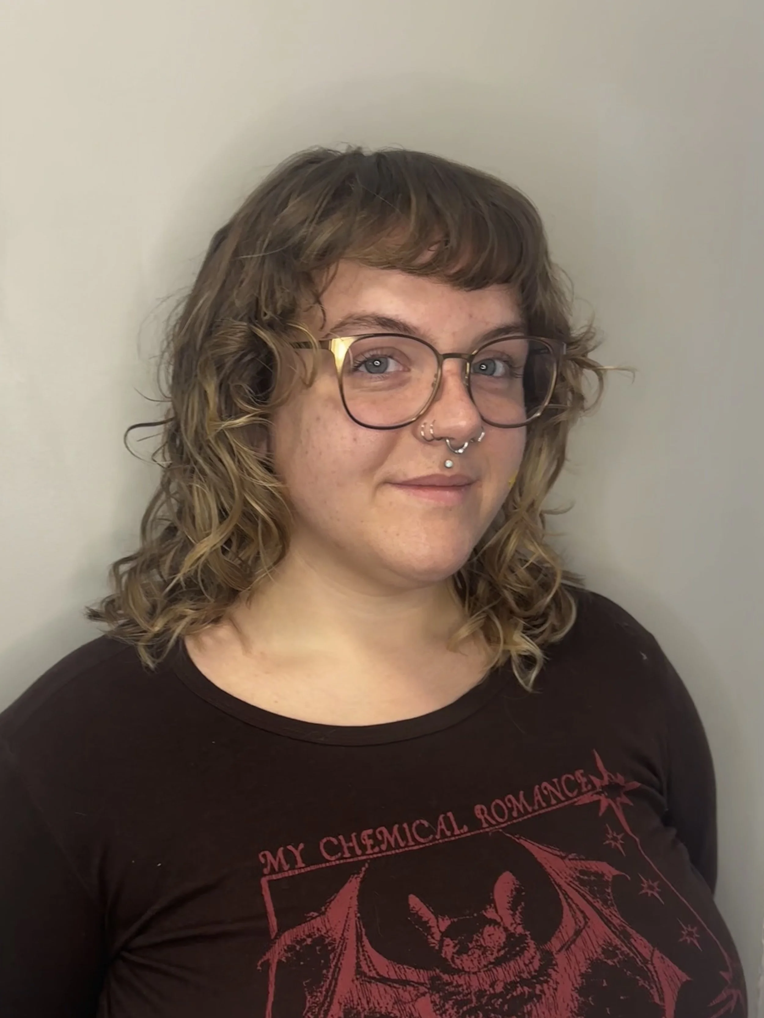 A woman with curly, shoulder-length hair, glasses, and a septum piercing, wearing a black illustrated shirt, standing against a plain light-colored wall.