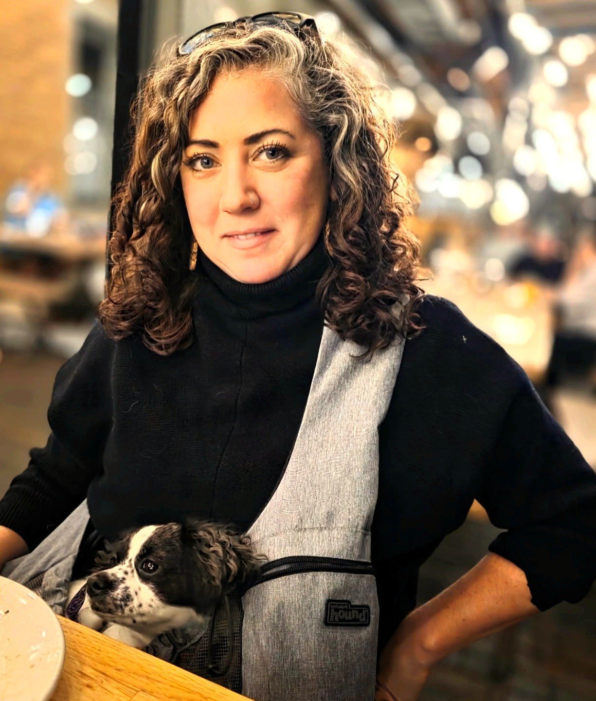 A woman with curly brown hair, wearing a black turtleneck and glasses on her head, sitting at a table with a small black and white puppy secured in a gray and black pet carrier. The background is blurred with warm lighting and out-of-focus people.