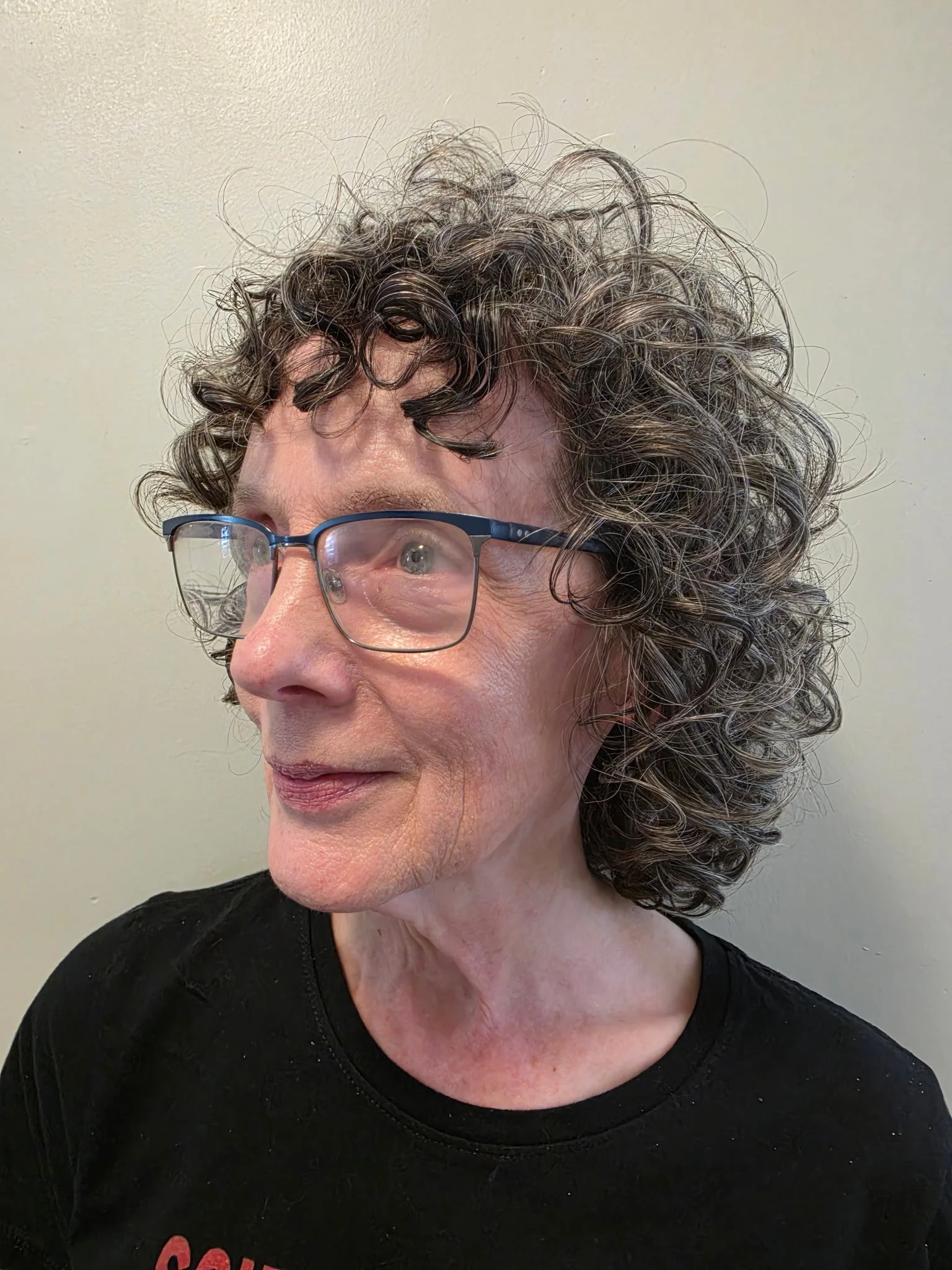 Close-up of a woman with curly gray hair, wearing glasses, and a black shirt, looking to the left against a plain background.