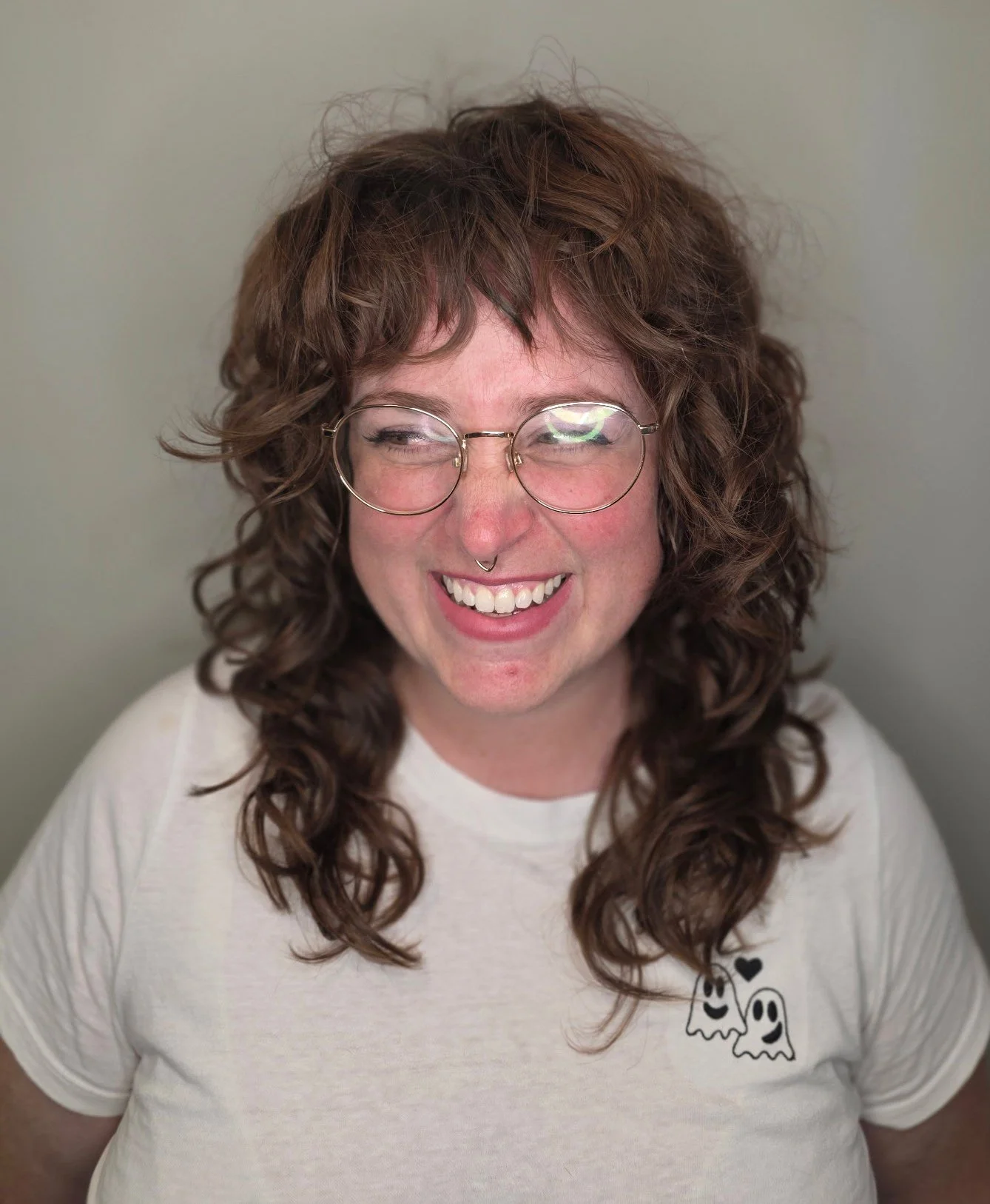 A young woman with curly brown hair, glasses, and a septum piercing, smiling and looking to her right, wearing a white T-shirt with a ghost and heart design.