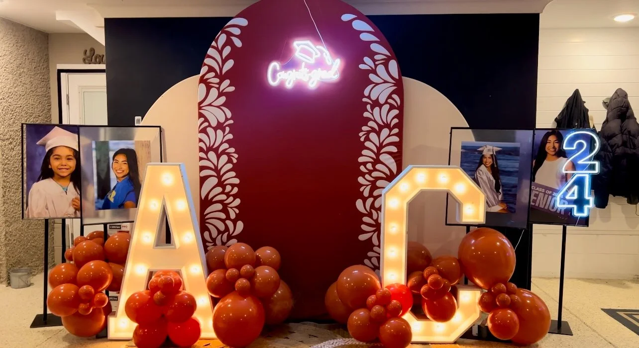A graduation celebration display with large illuminated letters A and C surrounded by orange balloons. Two framed photos of a young girl in a graduation cap and gown are on both sides, and a neon sign with a graduation cap and the words 'Congrats Grad' is at the top. Two projects with the number 2 and 4 are also present.