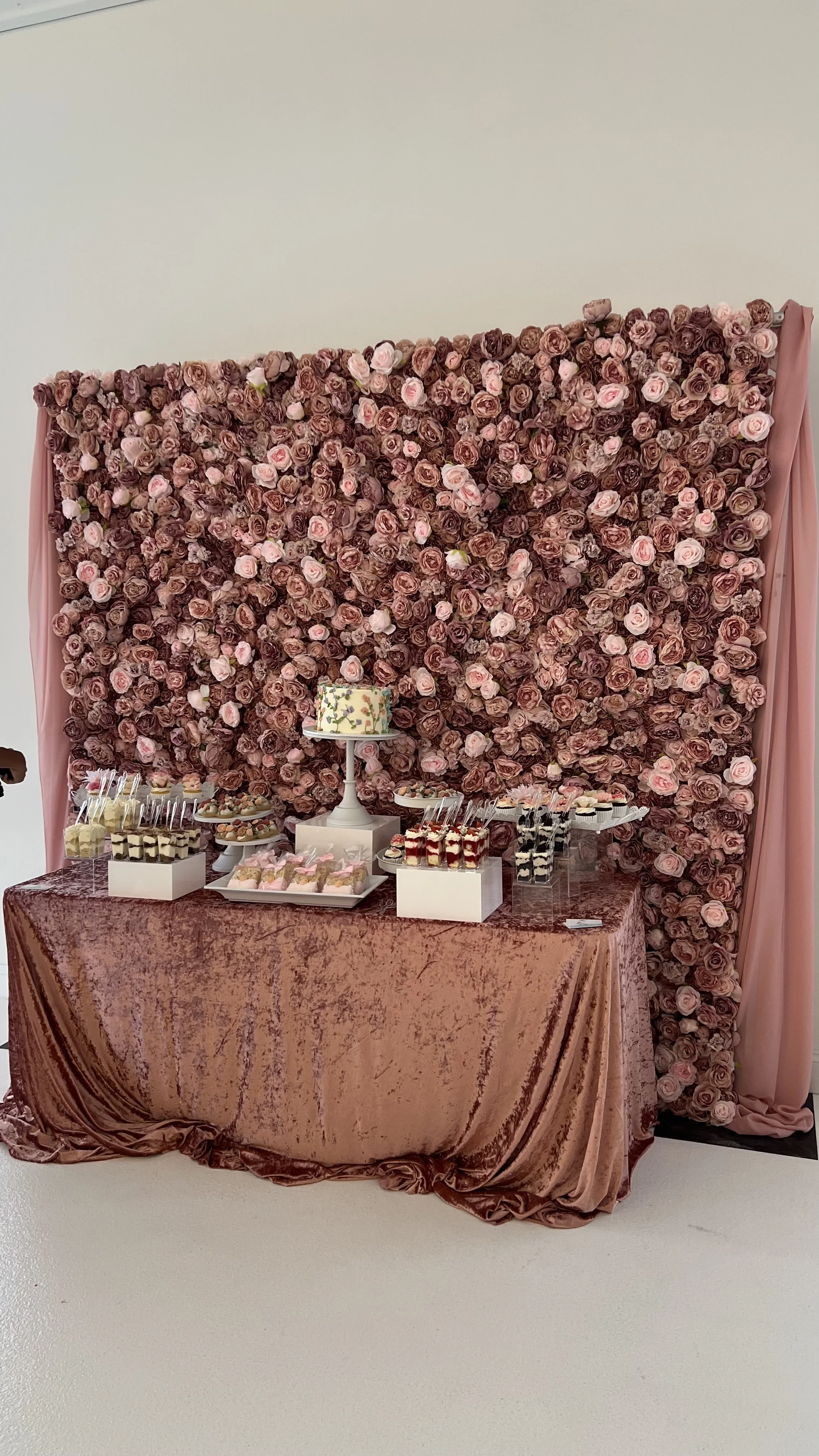Dessert table with a pink rose floral backdrop and pink velvet tablecloth, featuring a cake, cupcakes, and other sweets.