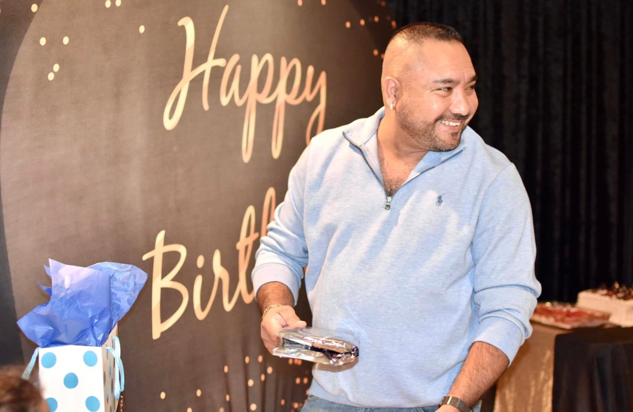 A man smiling at a birthday celebration, holding a wrapped gift, with a "Happy Birthday" backdrop and a gift bag nearby.