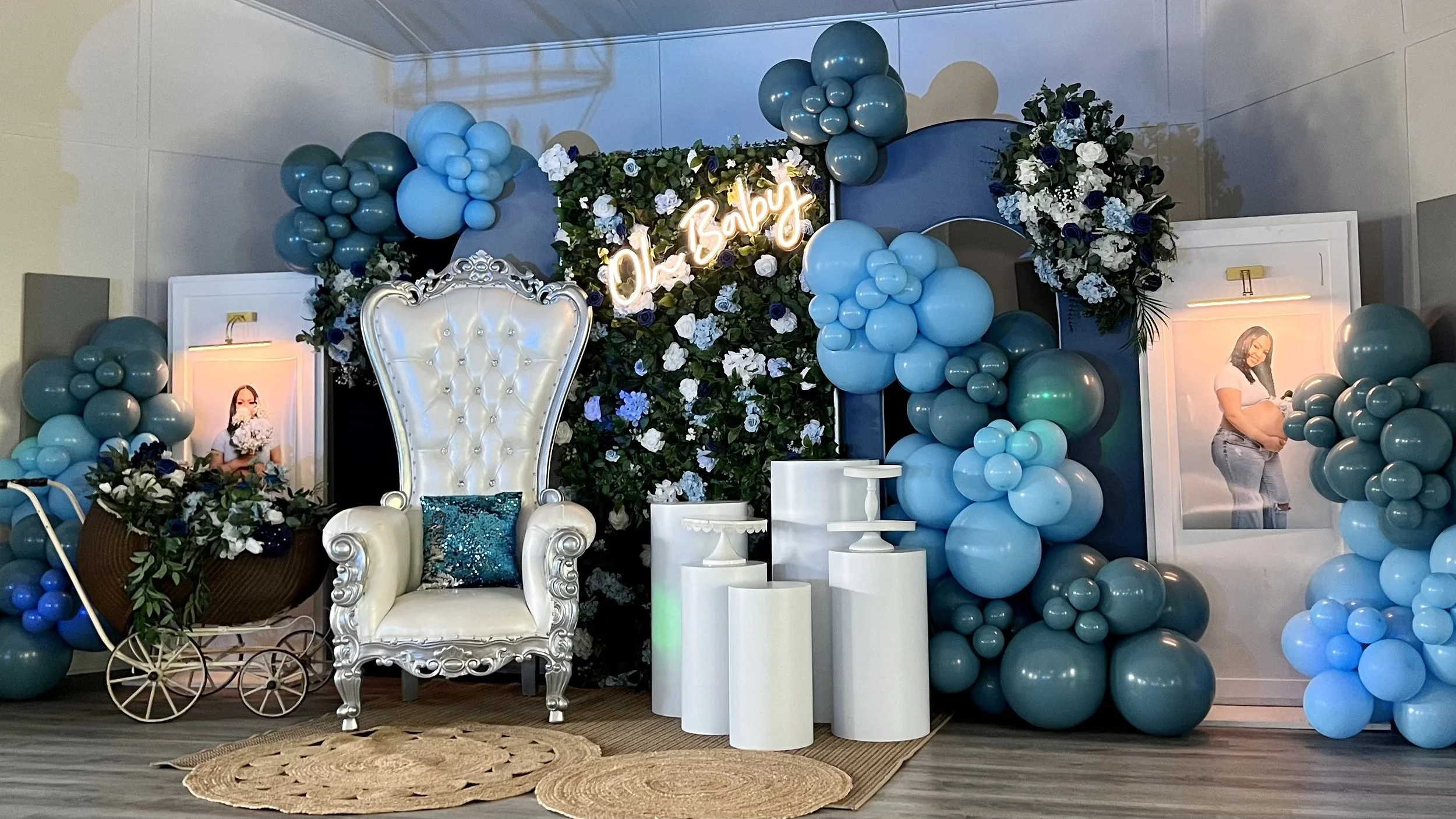 Decorative setup with a large white high-back throne, blue and white balloons, floral arrangements, a neon sign reading 'Ol Baby,' and framed pictures of a pregnant woman. The area includes small white tables, a brown wicker basket with flowers, and a beige woven rug on the floor.