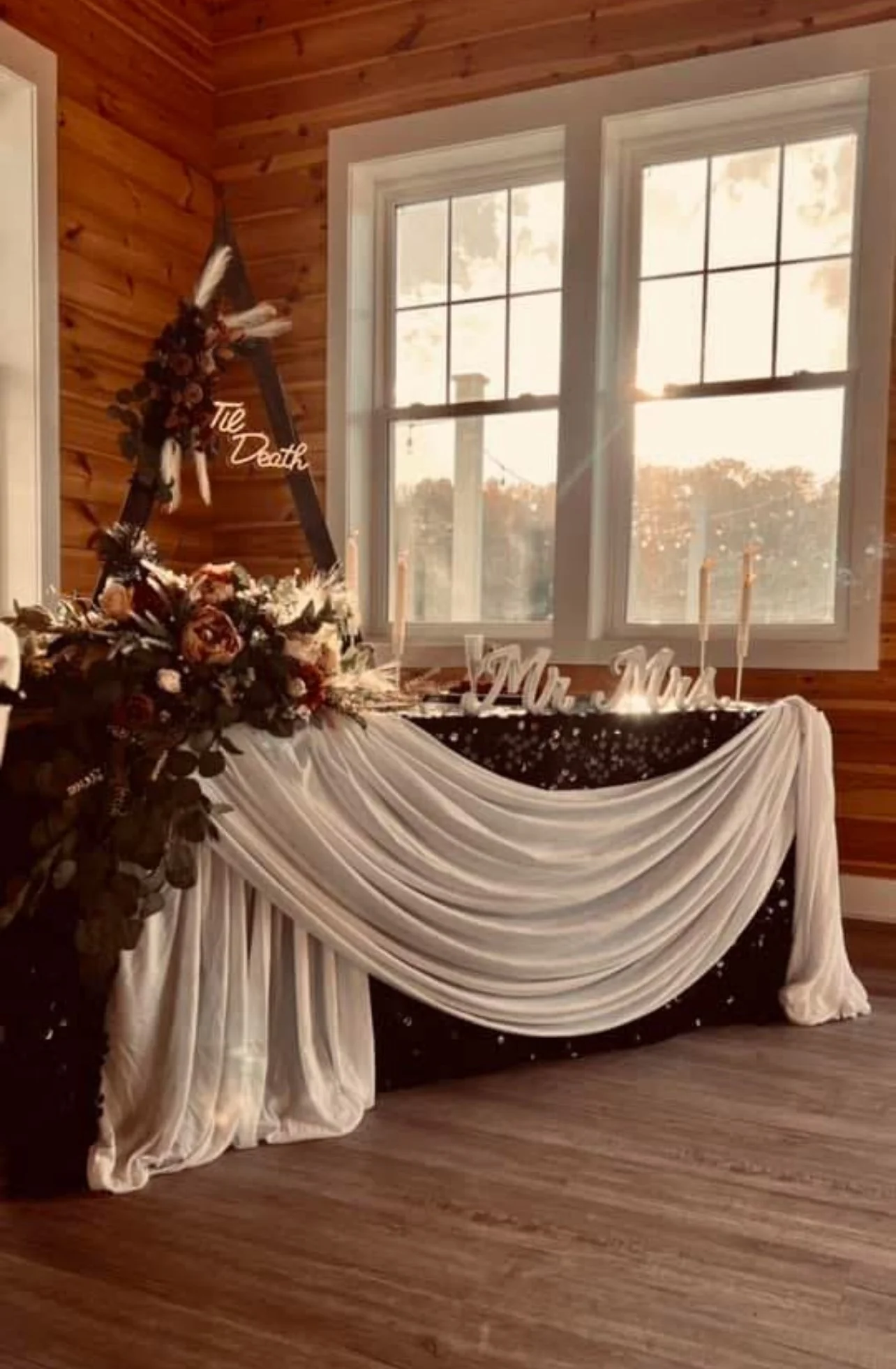 A decorated table with white drapery, floral arrangements, and candles, set inside a wooden room with large windows, likely for a wedding or celebration.