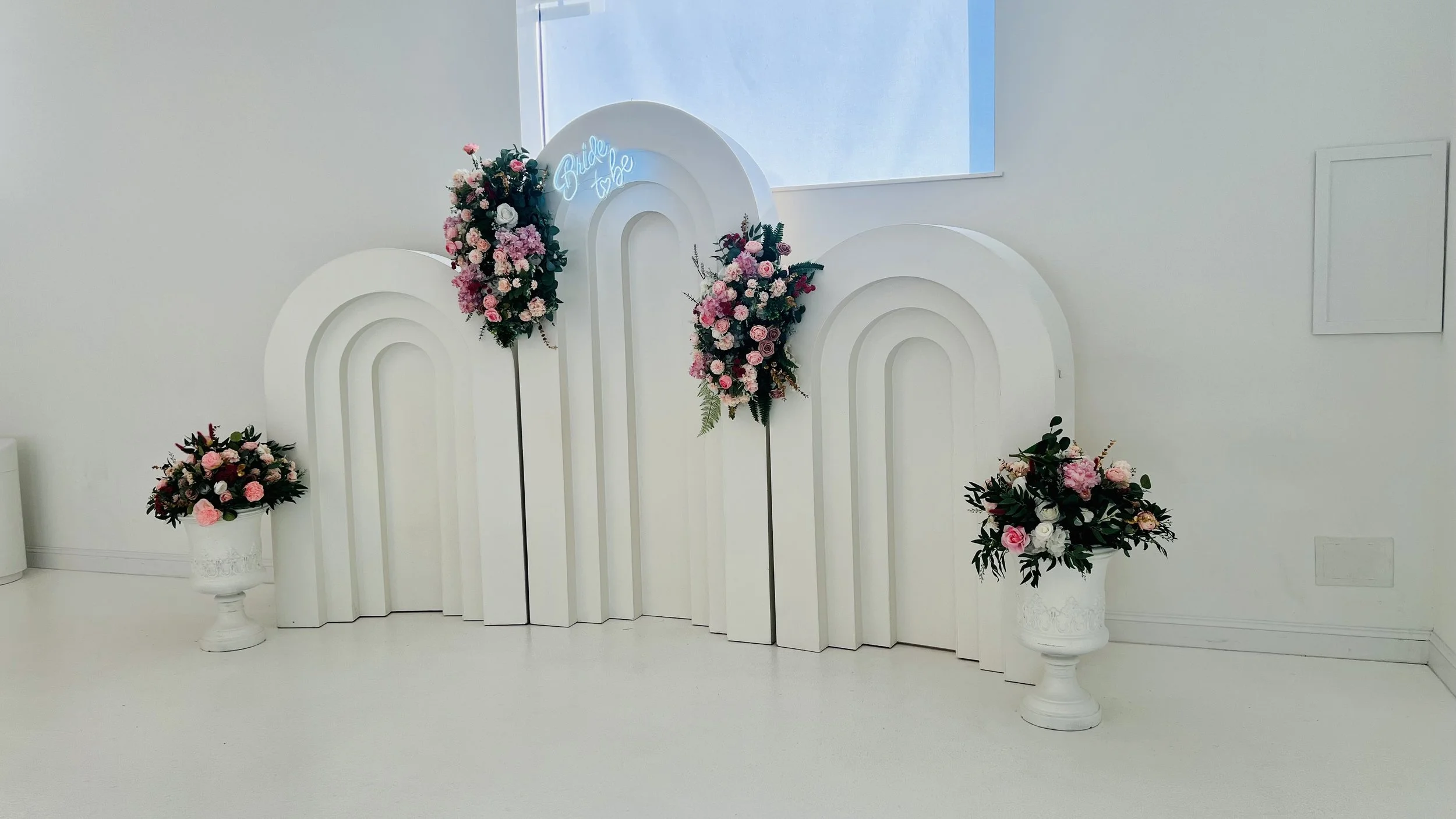 White decorative backdrop with pink and white floral arrangements, including roses and greenery, for a wedding or celebration.