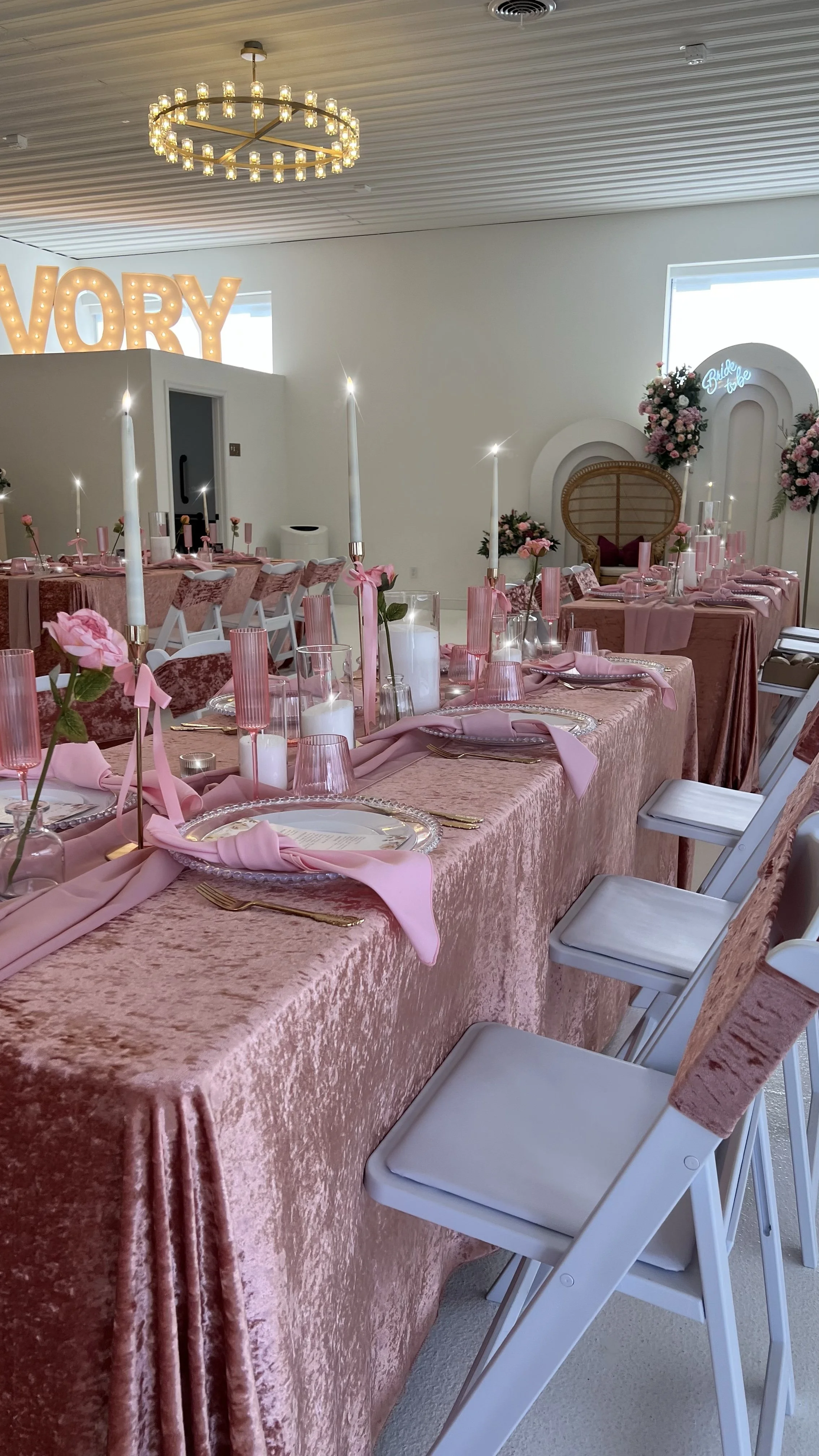 A pink-themed decorated event space with a long table set with pink cloth, pink napkins, gold cutlery, pink glassware, and floral centerpieces, with chairs placed around the table. Candles and tall candle holders are also on the table. In the background, there is a large illuminated sign spelling 'VORY' and decorative floral arrangements.