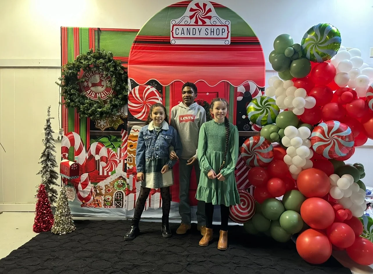 Three children standing in front of a festive holiday backdrop for a candy shop, decorated with large balloons, Christmas trees, and holiday decorations.