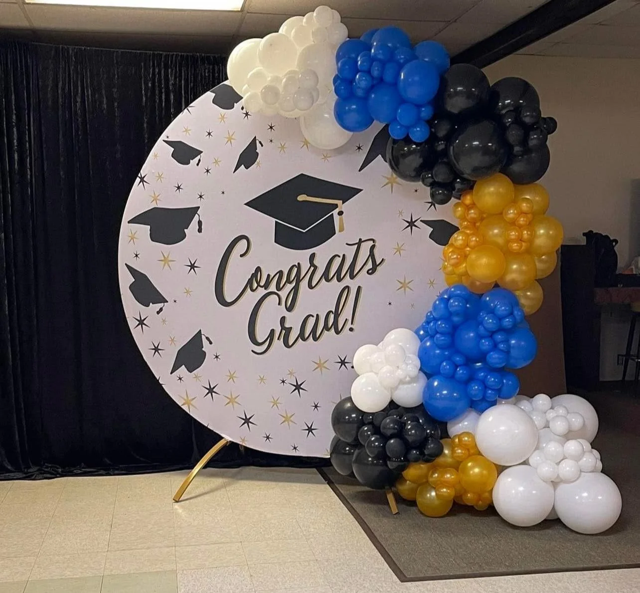 Graduation celebration display with a large round backdrop featuring black, white, and gold balloons arranged on the right, and black graduation cap silhouettes with stars and the text 'Congrats Grad!' in the center.