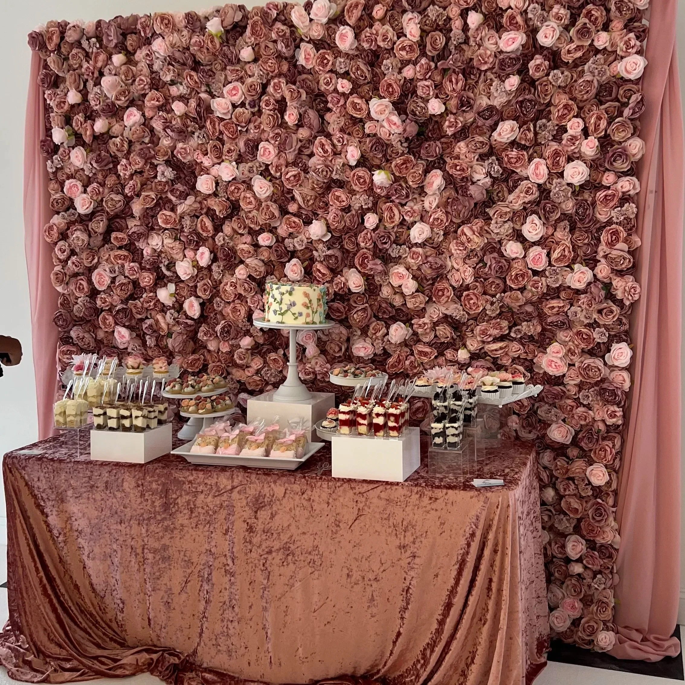 A dessert table with a pink velvet cloth in front of a large backdrop made of pink and purple roses. The table features an elegant white cake with floral decorations, cupcakes, and various small desserts displayed on white and clear stands.