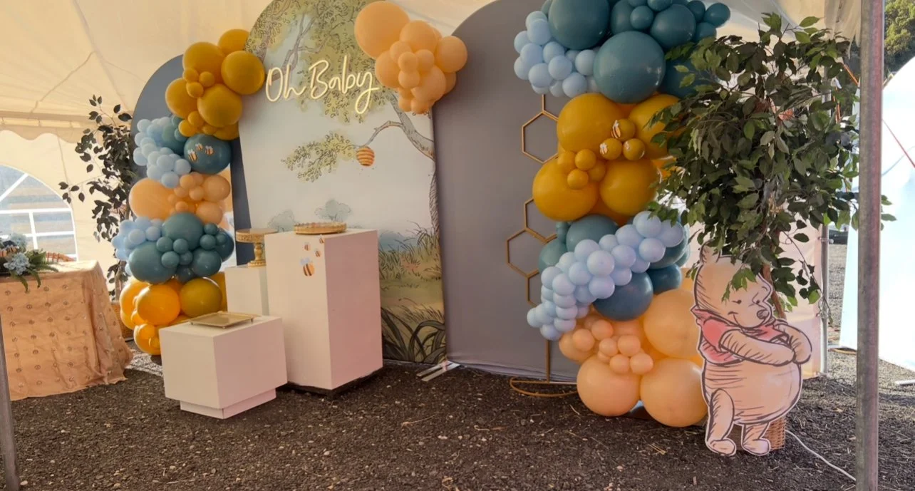 Decorative arch made of yellow, peach, and blue balloons with a baby-themed backdrop that says "Oh Baby". To the right, a cutout of a cartoon baby hugging its knees, next to a fake leafy plant. A table with pink cloth and flower arrangement is visible on the left.