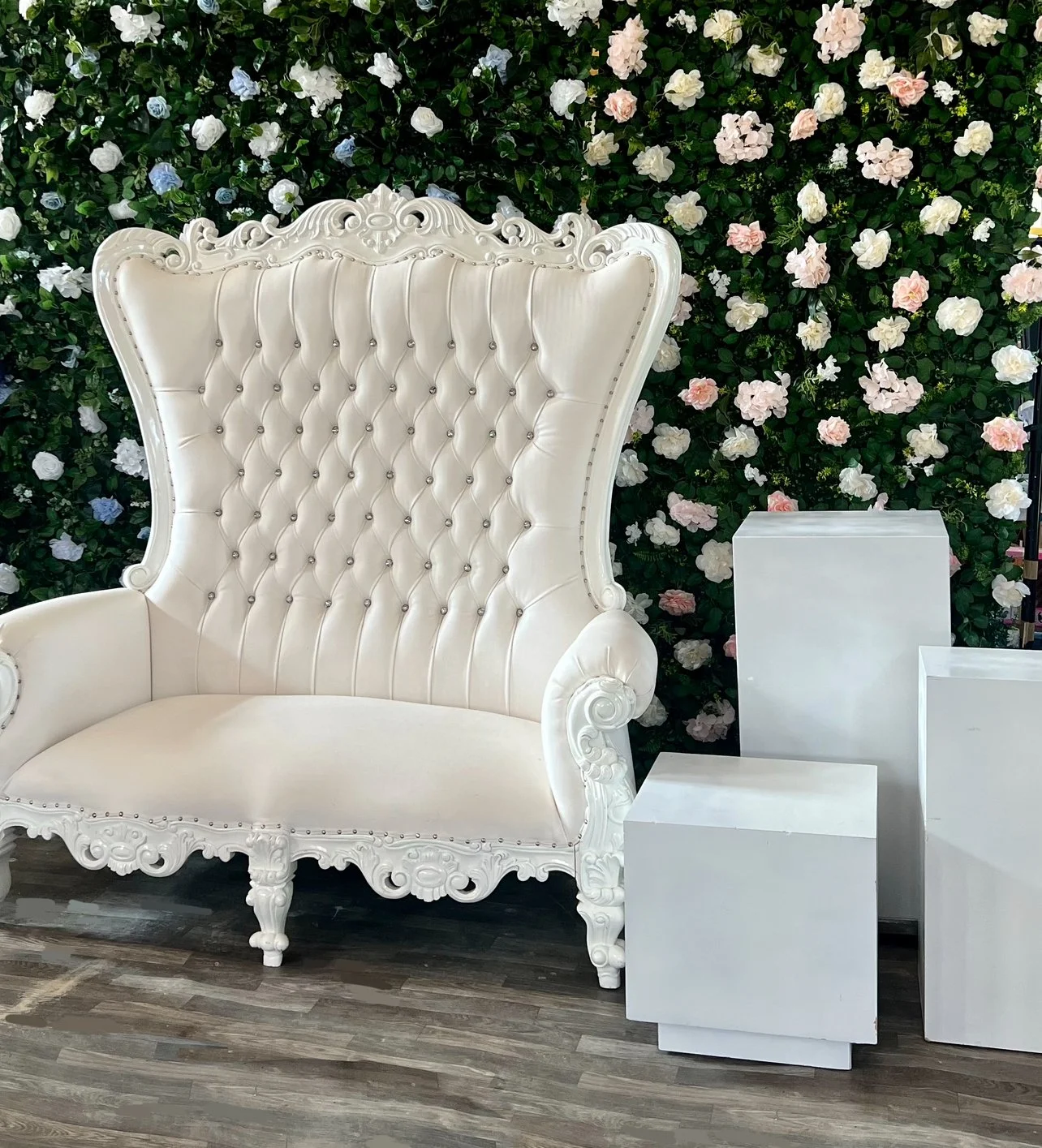A vintage white tufted armchair with carved wood detailing, placed in front of a lush green floral wall with pink and white flowers, and accompanied by several white rectangular pedestals on a wooden floor.