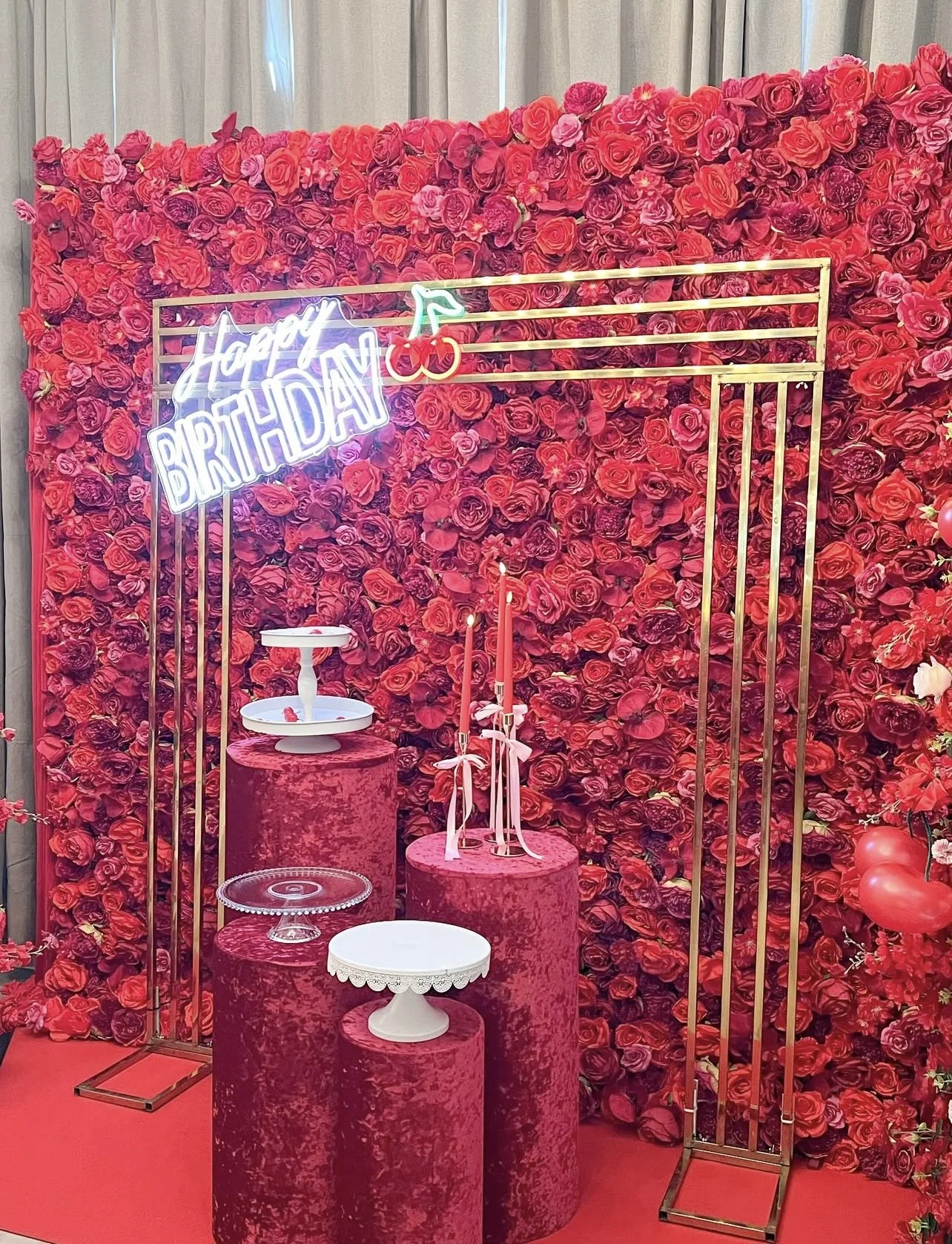 A birthday party setup with a red and pink floral backdrop, a neon sign that says 'Happy Birthday' with a cherry graphic, three pink pillars holding various decorative items including a tiered cake stand, a glass cake stand, and a lace-edged cake stand, with a red floor and gold metal frames on the sides.
