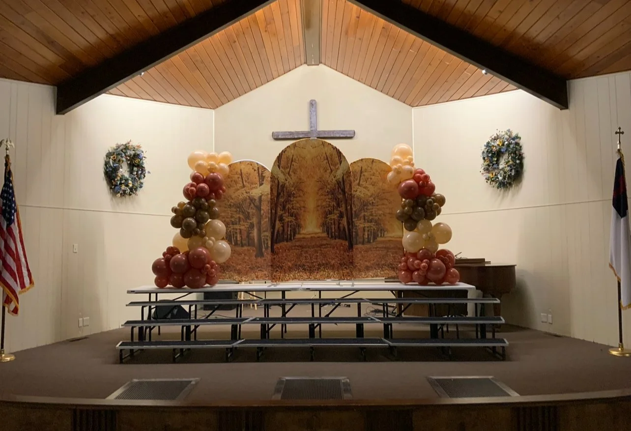 Interior view of a church stage decorated with a large backdrop of a forest pathway, balloon arches on either side in autumn colors, two wreaths on the walls, an American flag on the left, and a Christian flag on the right, with a cross mounted above the backdrop.