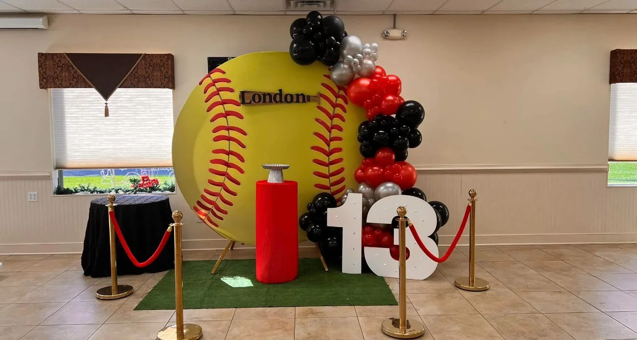 A baseball-themed birthday display with a large yellow baseball backdrop, balloons in red, black, and silver, and a white number 13. The word 'London' is on the baseball backdrop. The display is surrounded by red ropes on gold stanchions and is set on a small green mat inside a room with windows.