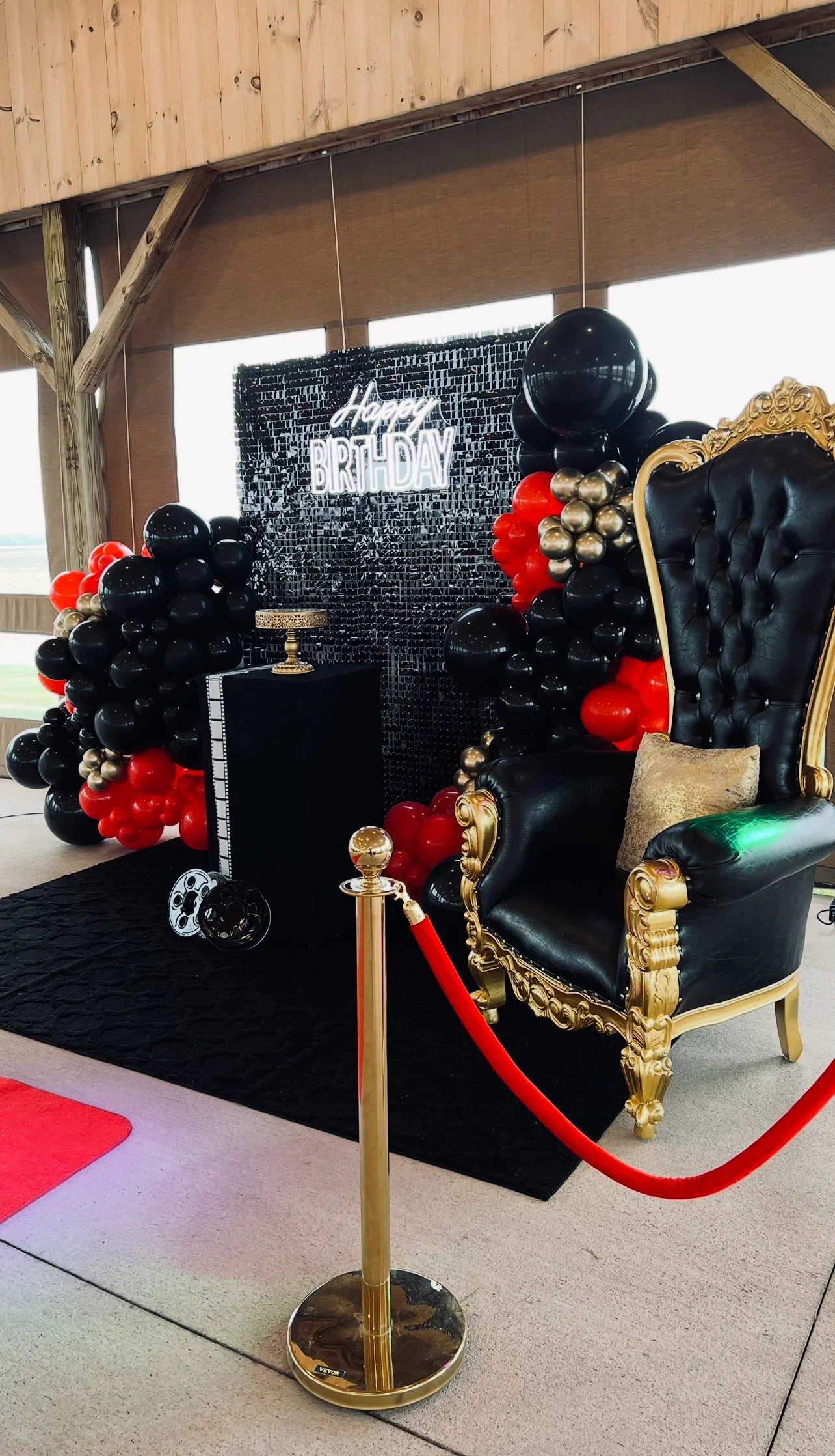 Birthday celebration setup with a black and gold throne, a backdrop with balloons in black, red, and gold, and a neon sign that reads 'Happy Birthday'.