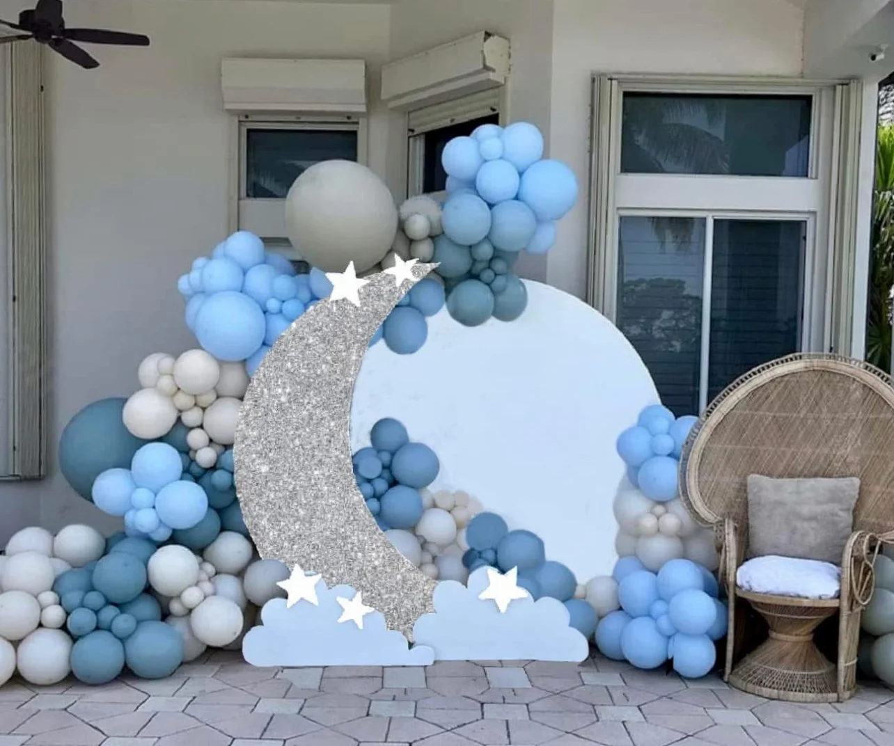 Decorative setup with a large white circular backdrop surrounded by blue and white balloons, featuring a glittery silver moon with stars and clouds, placed on a tiled outdoor patio near a wicker chair with a cushion.