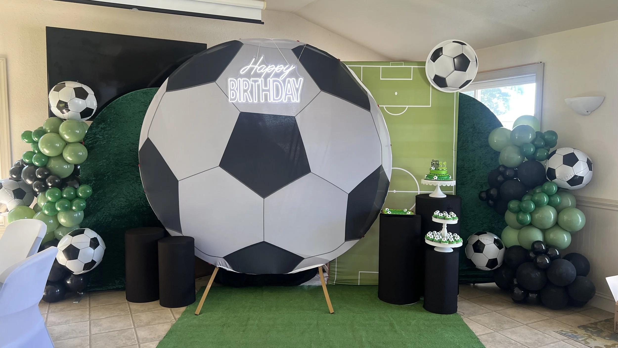 Soccer-themed birthday party setup with large soccer ball decoration, balloon arrangements of green, black, and white, and cake stands with soccer-themed cupcakes, on a green floor mat.