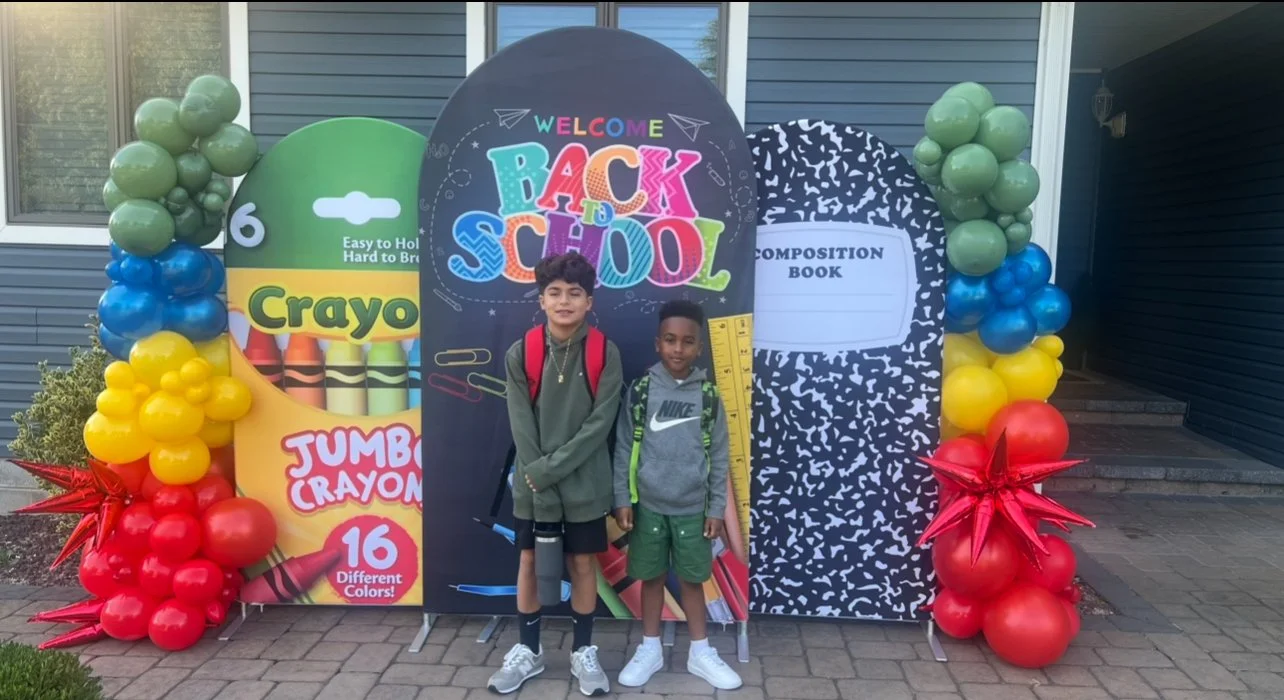 Two boys standing in front of a colorful back-to-school display with balloons, a crayon-themed poster, and a composition book backdrop, celebrating the start of the school year.