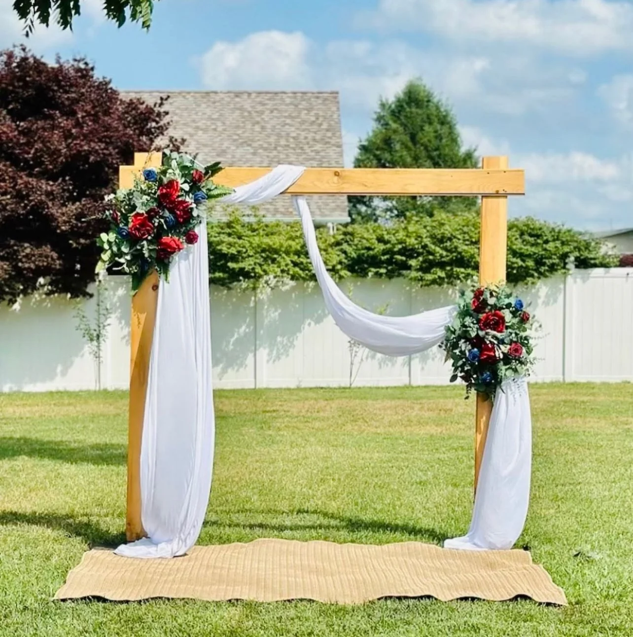 Wooden wedding arch decorated with white fabric and bouquets of red and blue flowers, set up outdoors on grass with a white fence and trees in the background.