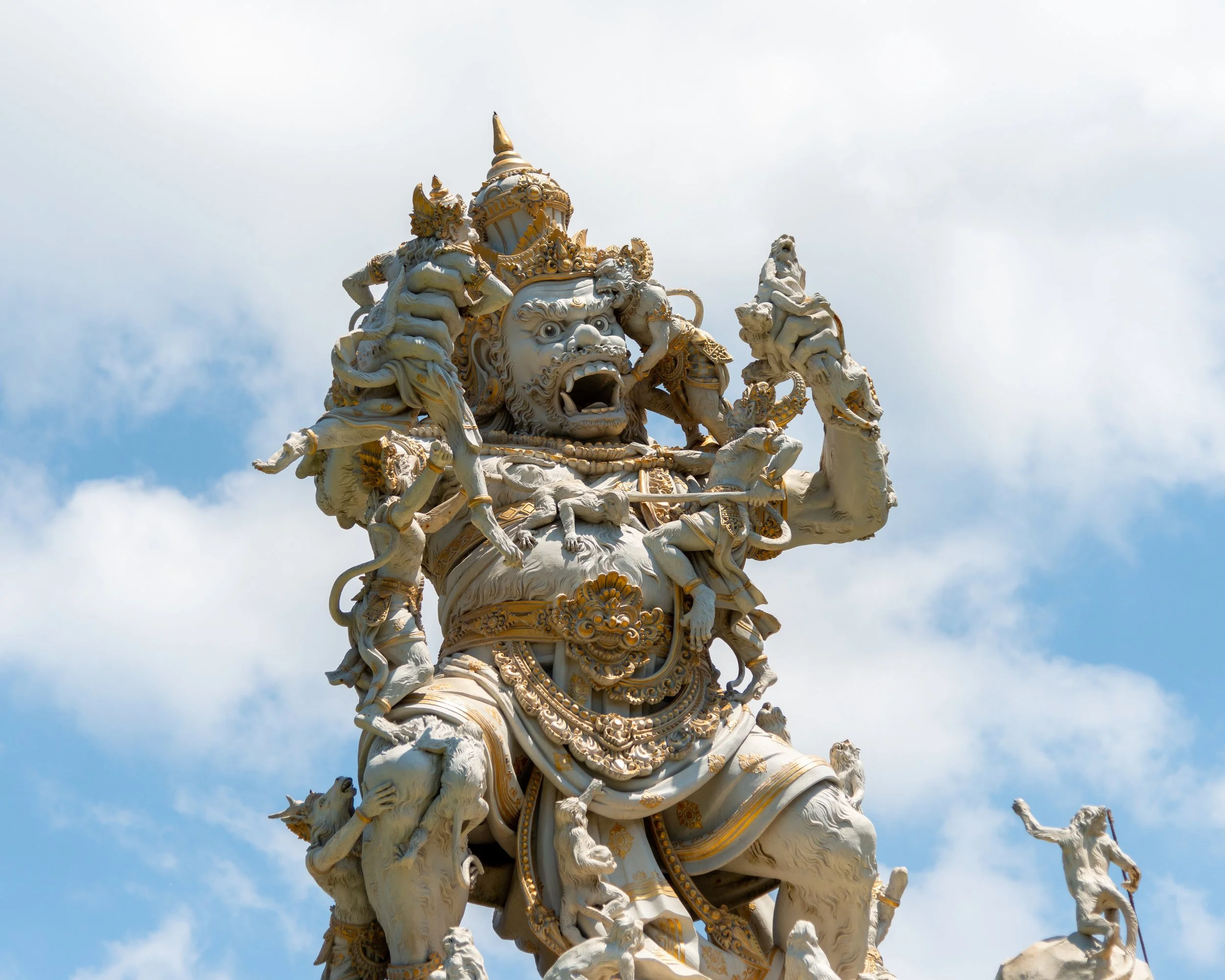 Ornate white and gold sculpture of a fierce mythological figure with multiple heads and arms against a cloudy sky.