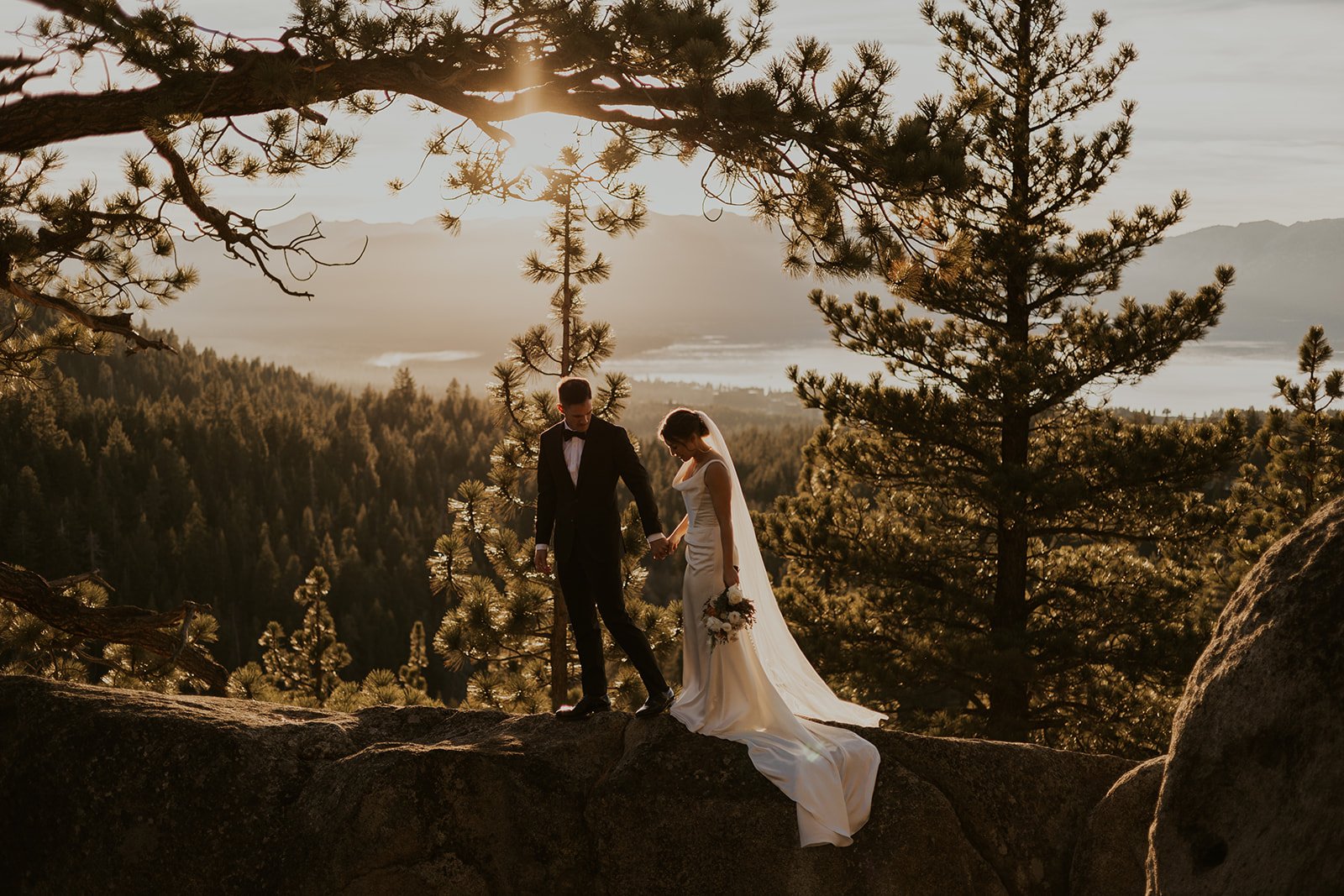 A groom holding his bride's hand and guiding her across a rocky ledge while they look down. The bride is in her wedding gown and long white veil and holding her bouquet in the other hand. The sun is setting over Lake Tahoe in the background.